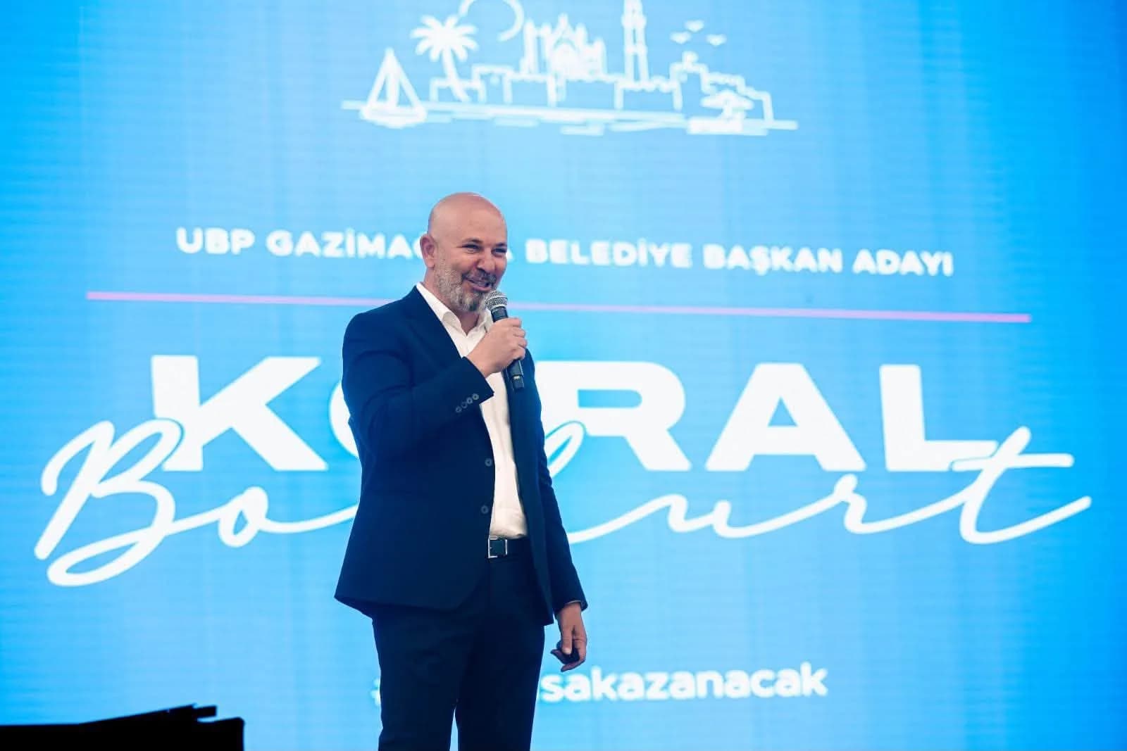 Koral Bozkurt, sahnede gülümseyerek konuşurken, arkasında kampanya ekranı